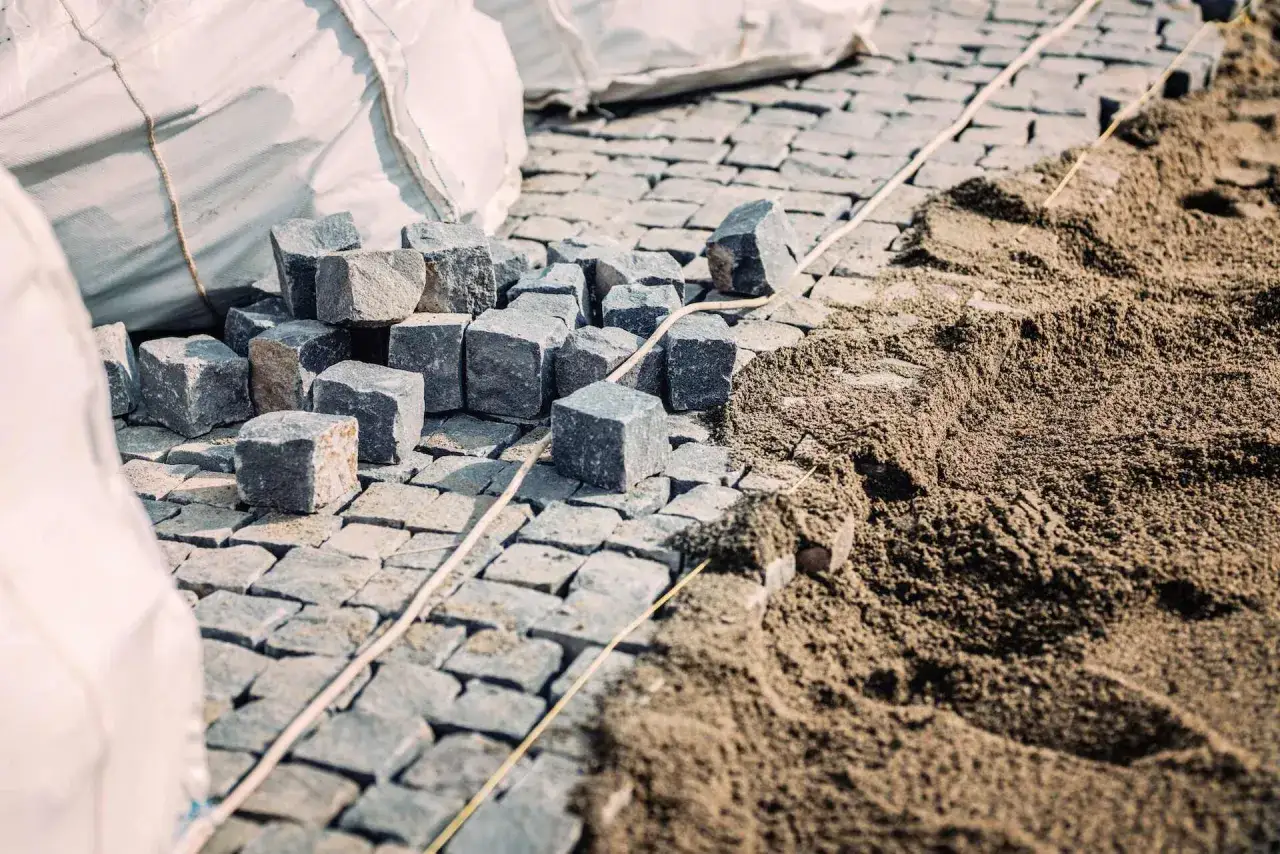Kostka brukowa i piasek na budowie. Widać stos kostki i przygotowany pod nią piasek, który posłuży jako podsypka pod kostkę. Ile cm piasku? To zależy od projektu.