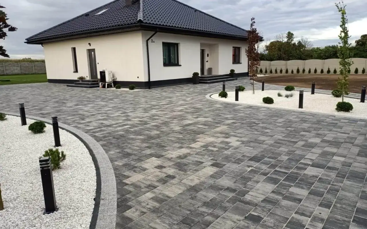 Nowoczesna kostka przed domem tworzy elegancką nawierzchnię. Wokół domu biały żwir, niskie krzewy i stylowe lampy.