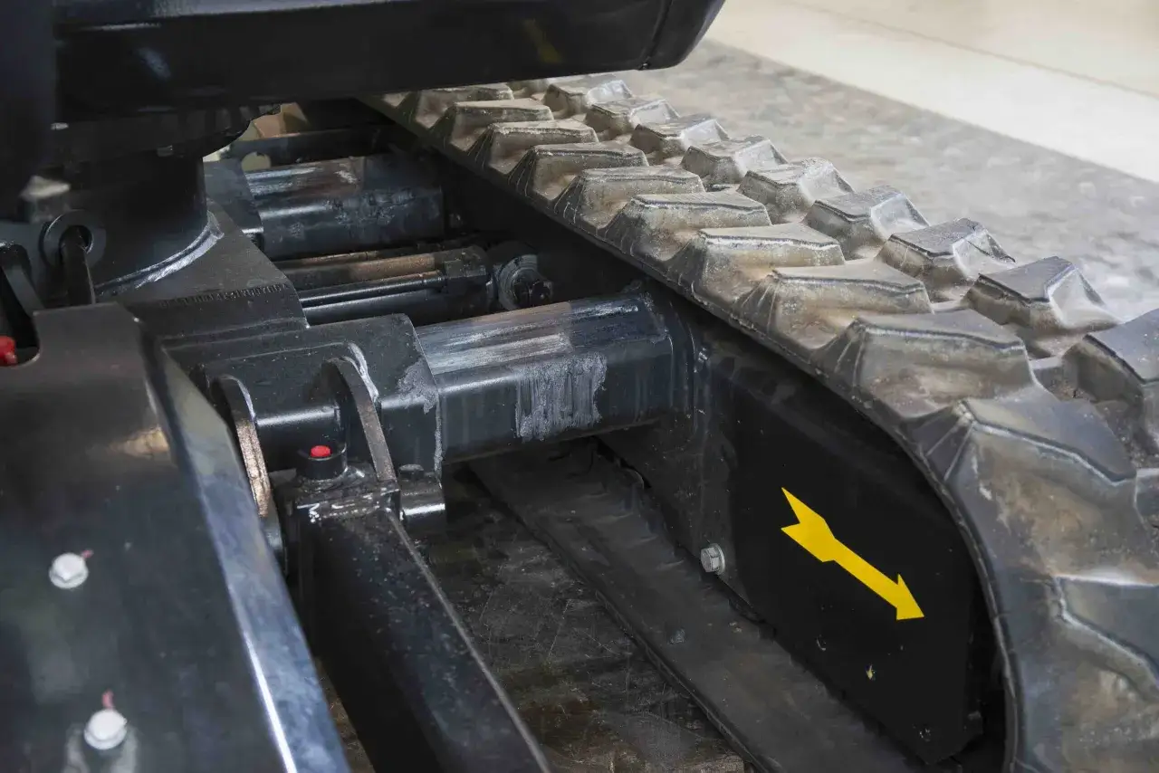 Zbliżenie na mechanizm minikoparki, pokazujące jak założyć gąsienice. Widoczna gumowa gąsienica i elementy podwozia.