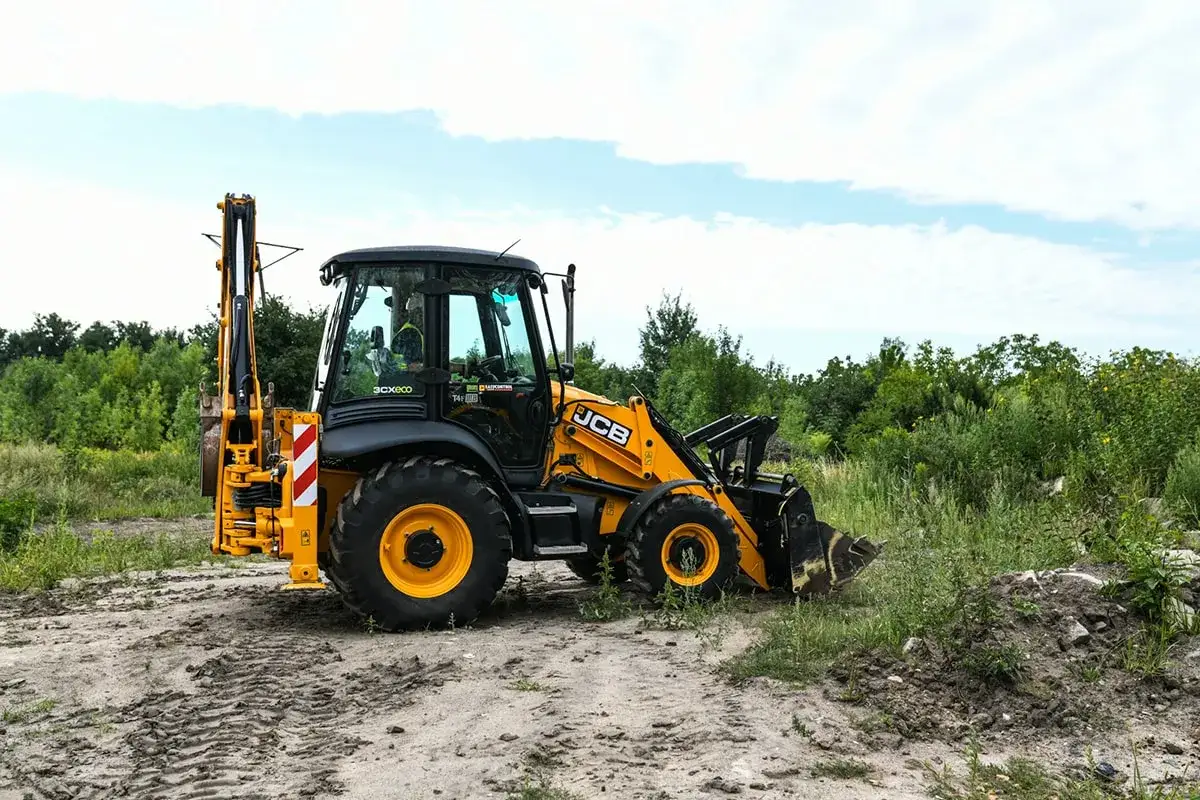 Żółta koparko-ładowarka JCB z uprawnieniami do pracy na budowie, gotowa do akcji na tle zieleni.